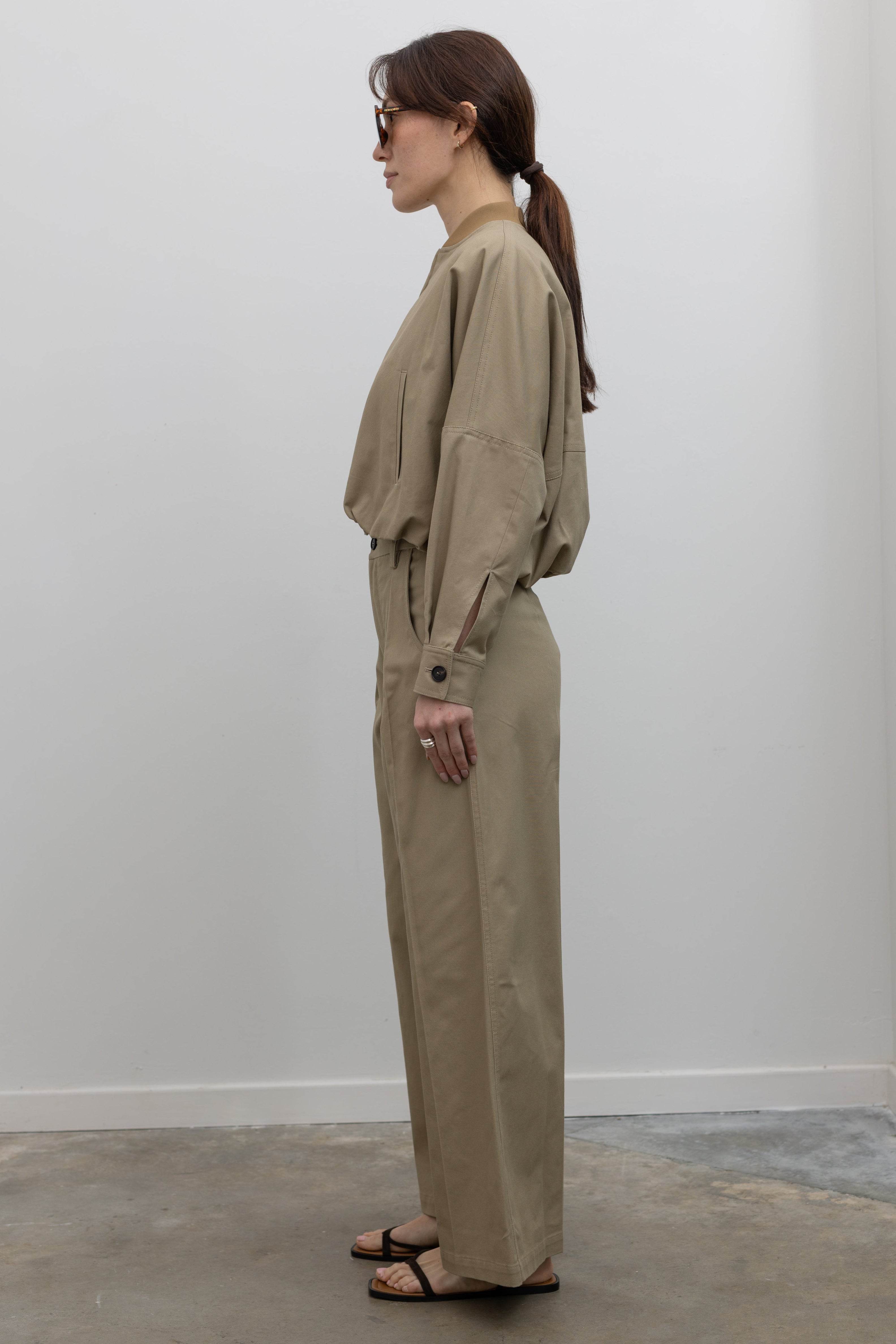 Side view of Woman standing wearing Spring fashion in a pair of Tela Rubicon Sand Cotton Gabardine Trousers and cropped jacket with sunglasses and sandals at Alas Eius