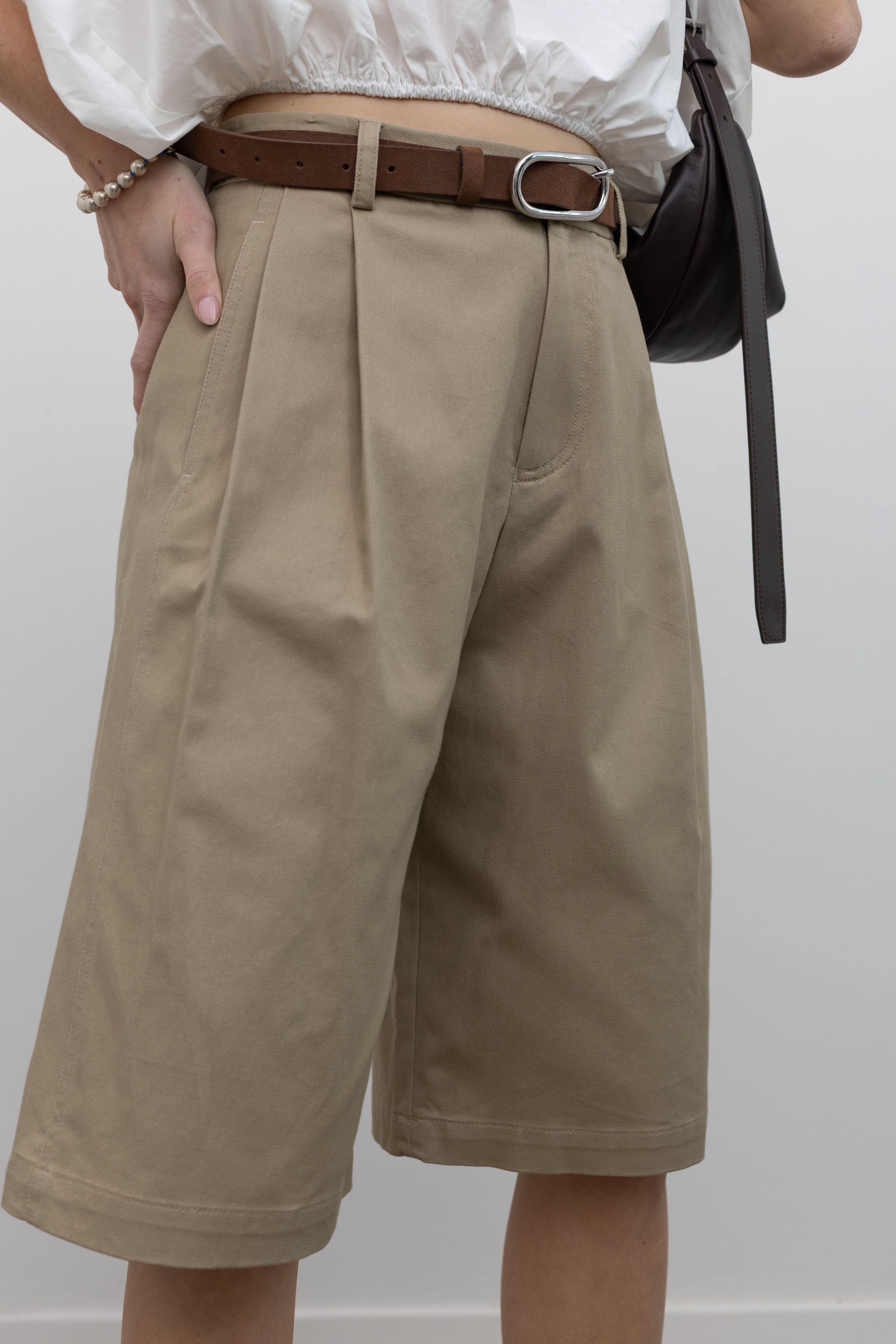 Close-up 3/4 view of a woman wearing cool women's street style fashion in the Tela sand cotton gabardine Bermuda long short featuring a front pleat and side seam pockets paired with a classic brown leather belt, a crisp white top, a St. Agni leather bag, and white leather pointed toe kitten heels at an Alas Eius store. 