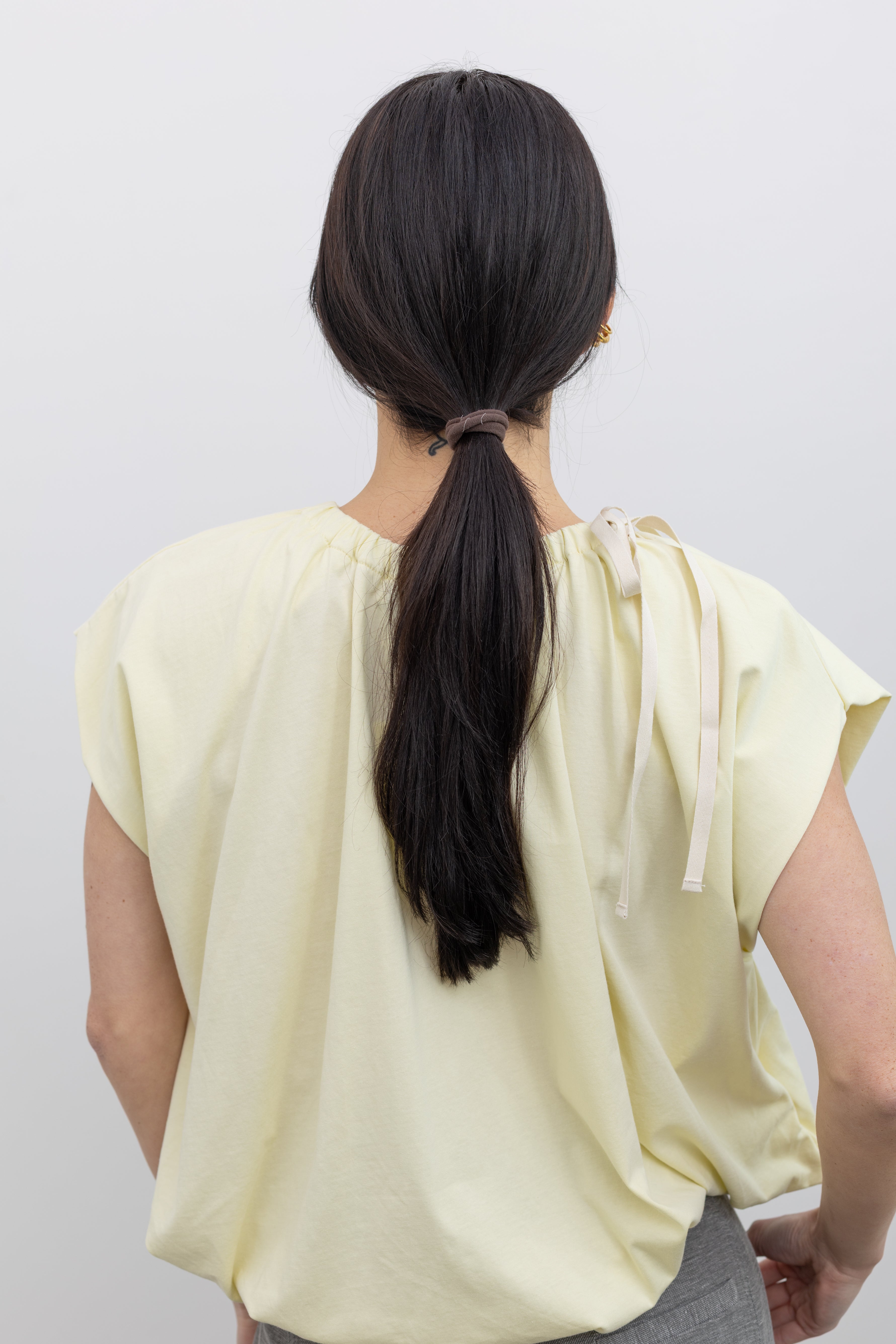 Close-up back view of a woman wearing spring fashion in a Tela pastel yellow gathered neck bubble hem short sleeve top at Alas Eius.
