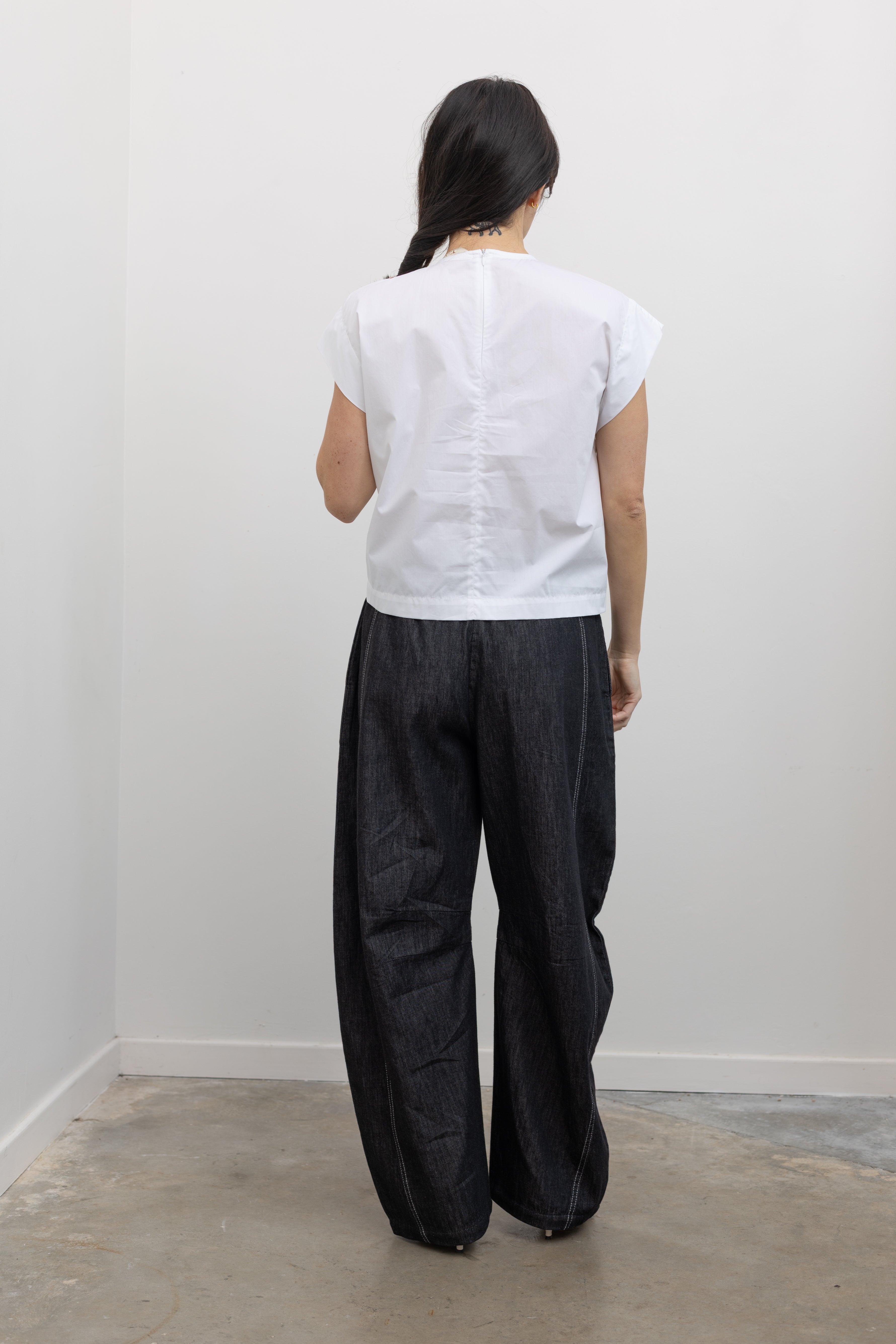 Full length back view of a woman wearing modern minimalist spring fashion in a Tela brand white Livia shirt featuring a front pleat that creates an intentional asymmetrical drape, crew neckline and relaxed cap sleeves paired with oversized baggy barrel leg pants at at an Alas Eius store. 