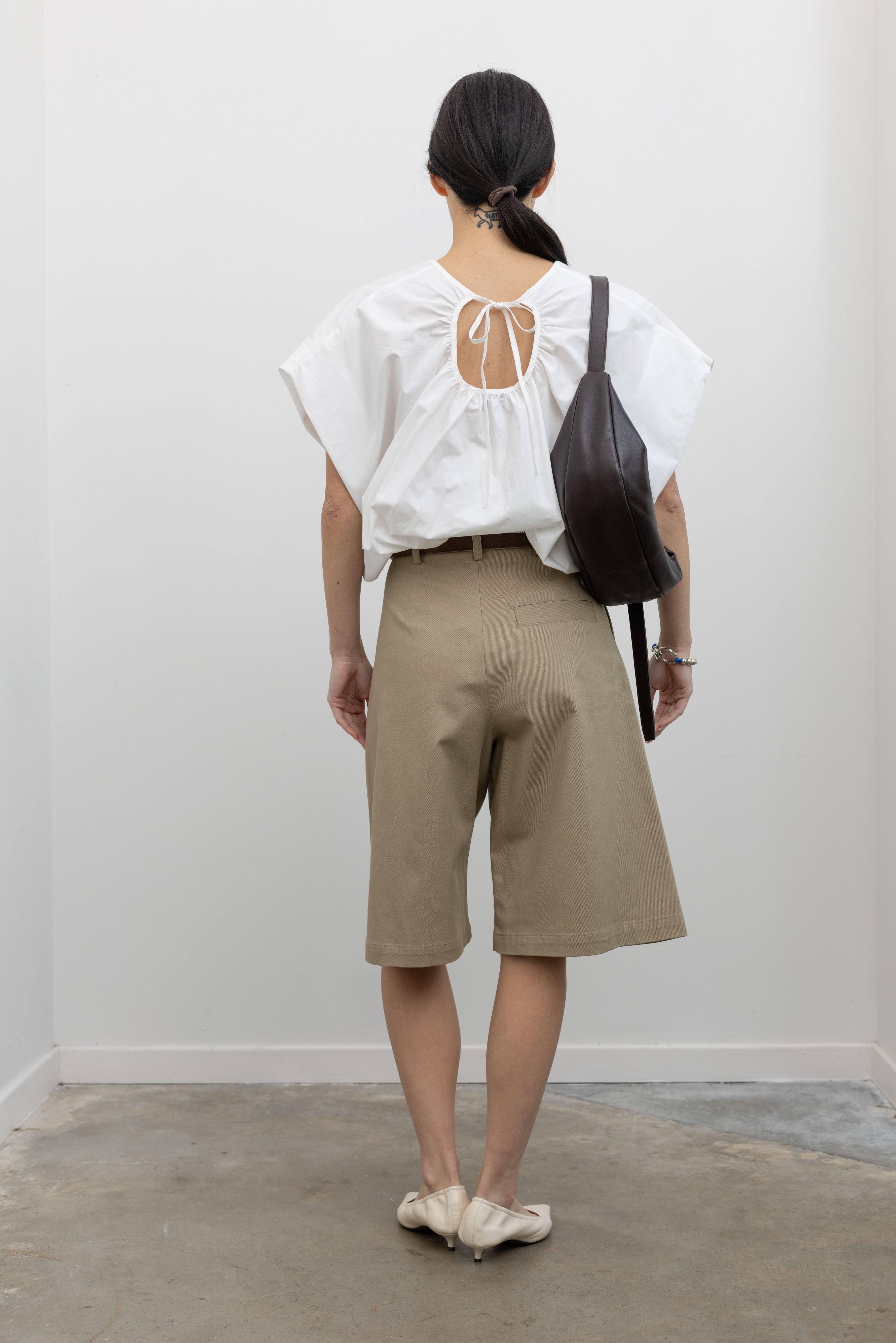 Full length back view of a woman posing wearing an elevated and chic outfit in the Tela sand cotton gabardine Bermuda long short featuring a front pleat and side seam pockets paired with a classic brown leather belt, a crisp white top, and white leather pointed toe kitten heels at an Alas Eius store. 