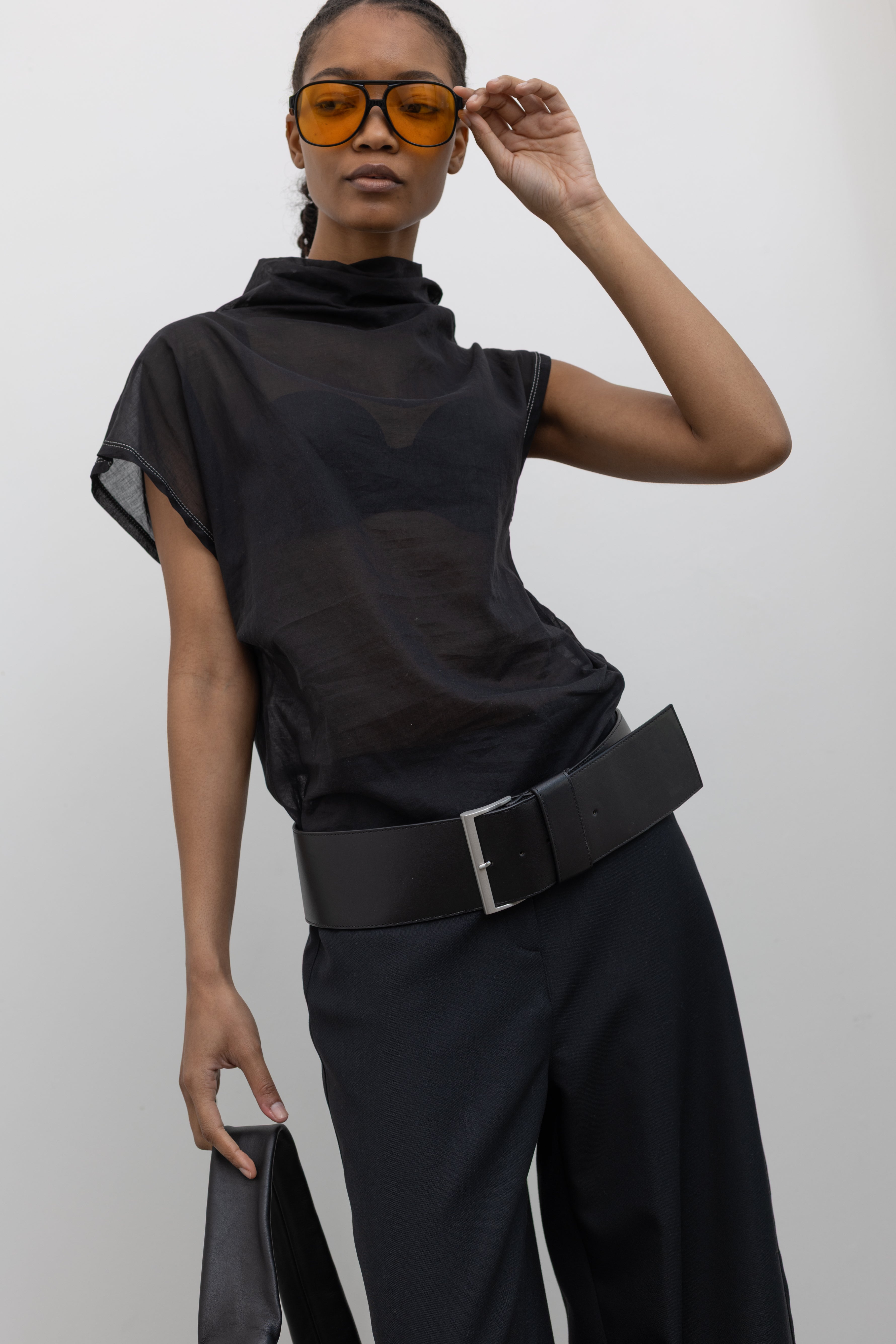 Close-up front view of a woman posing wearing a unique Tela brand semi-sheer lightweight washed muslin black top featuring a soft draped asymmetrical neckline paired with a classic wide black leather belt on black wide leg trousers at an Alas Eius store. 