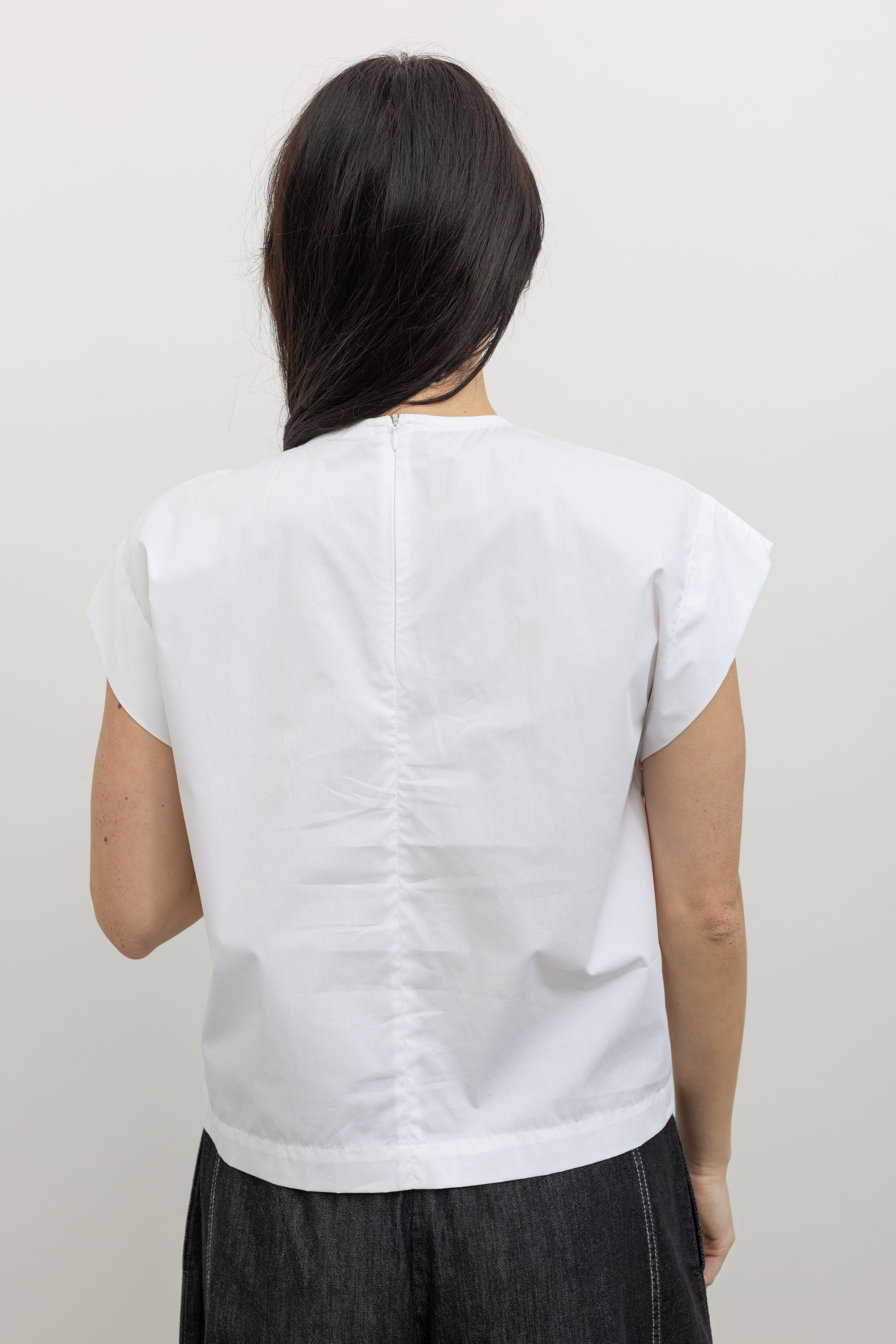 Close-up back view of a woman wearing an elevated classic top in a Tela brand white Livia shirt featuring a front pleat that creates an intentional asymmetrical draped panel, crew neckline and relaxed cap sleeves paired with oversized baggy barrel leg pants at at an Alas Eius store. 
