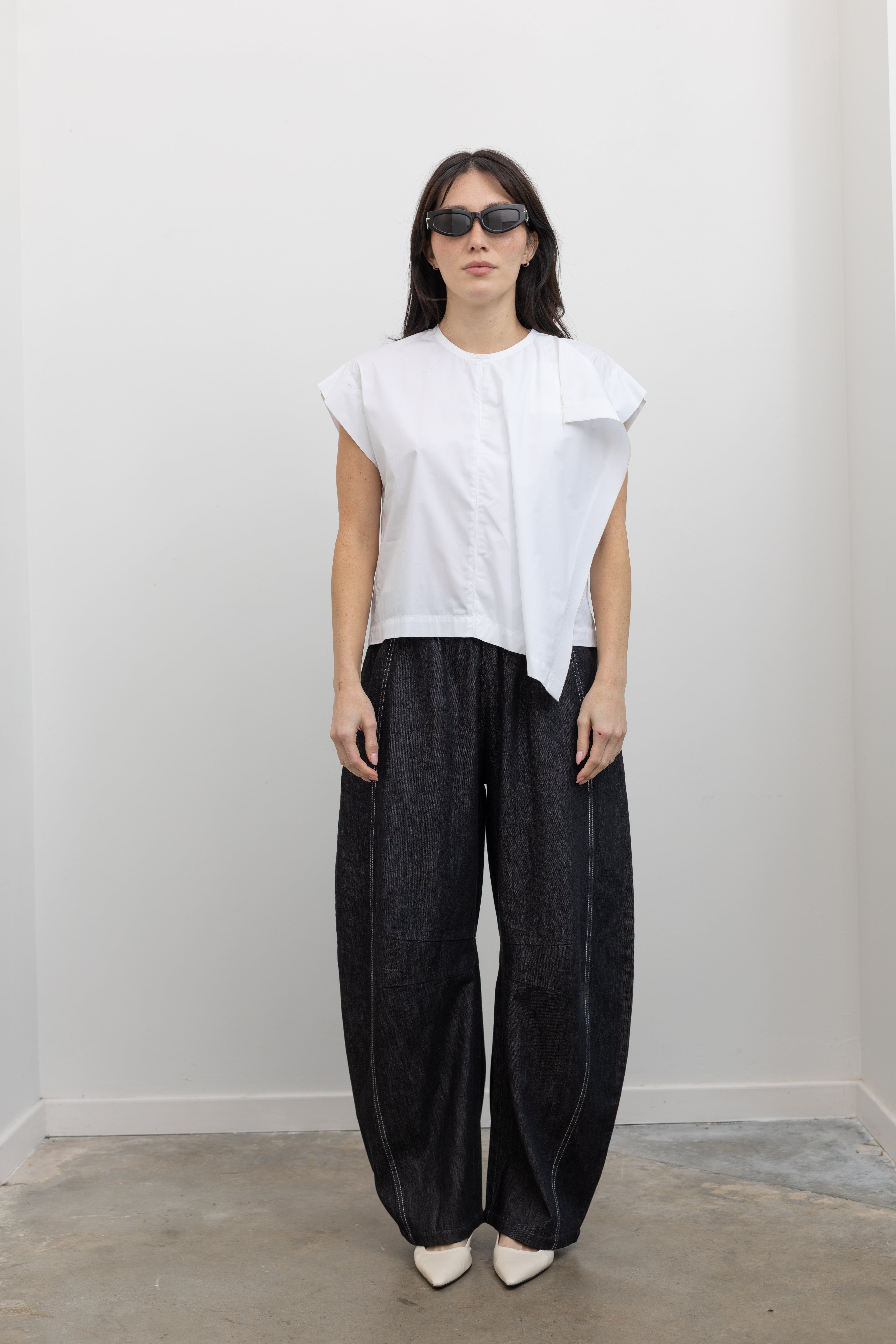 Full length front view of a woman wearing elevated spring fashion in a Tela brand white Livia shirt featuring a front pleat that creates an intentional asymmetrical drape, crew neckline and relaxed cap sleeves paired with oversized baggy barrel leg pants at at an Alas Eius store. 