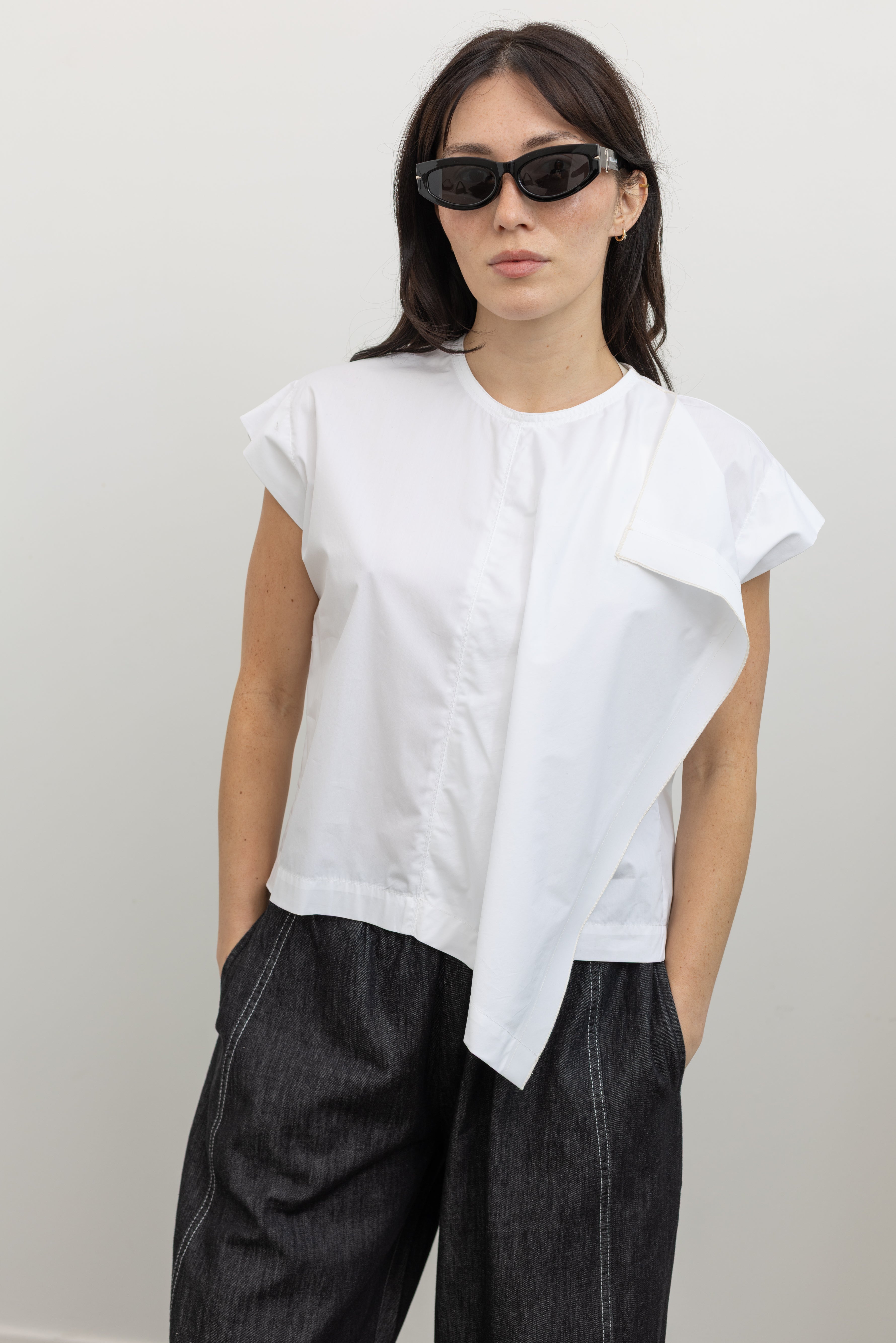 Close-up front view of a woman wearing elevated spring fashion in a Tela brand white Livia shirt featuring a front pleat that creates an intentional asymmetrical drape, crew neckline and relaxed cap sleeves at an Alas Eius store. 
