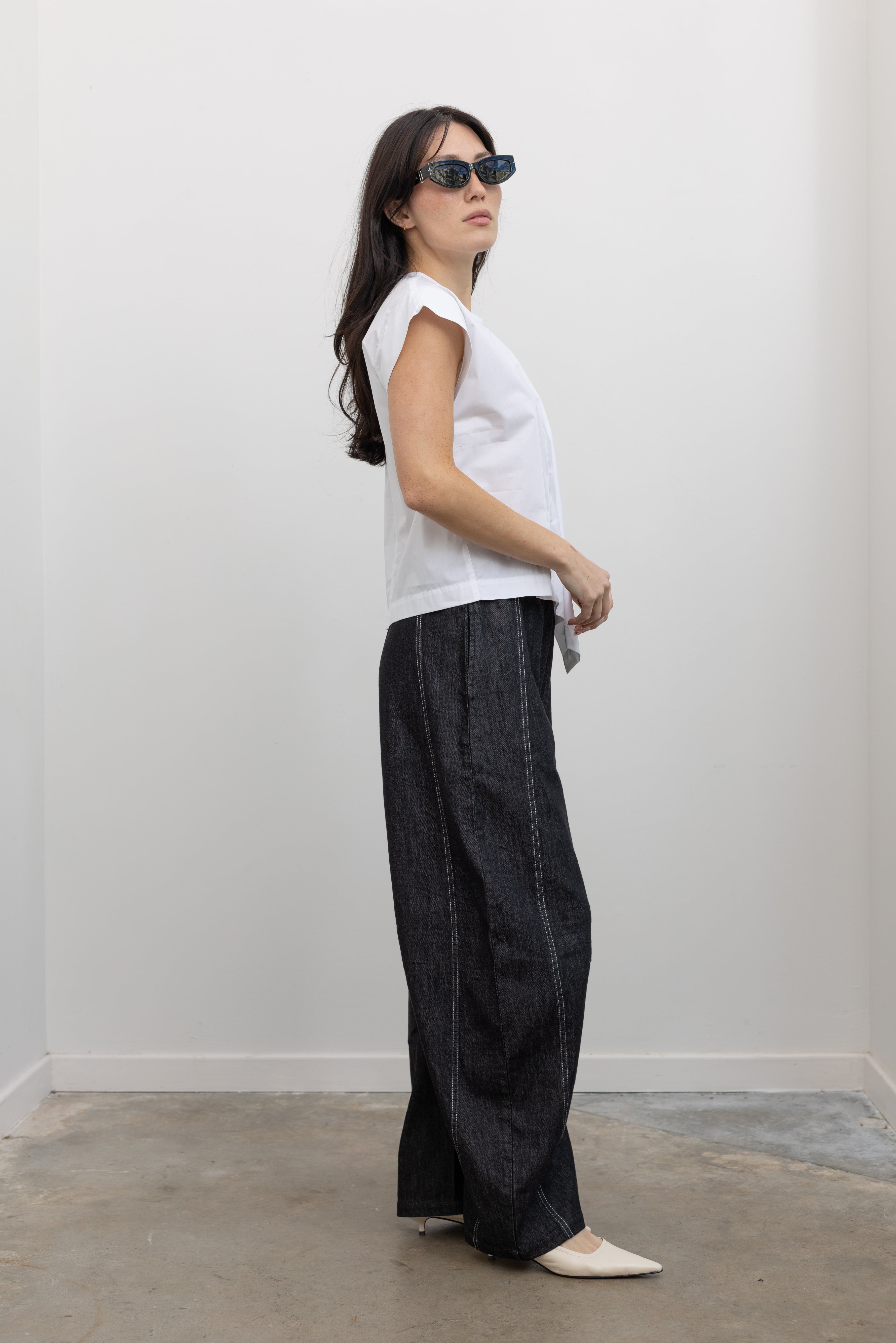 Full length side view of a woman wearing modern minimalist spring fashion in a Tela brand white Livia shirt featuring a front pleat that creates an intentional asymmetrical drape, crew neckline and relaxed cap sleeves paired with oversized baggy barrel leg pants at at an Alas Eius store. 