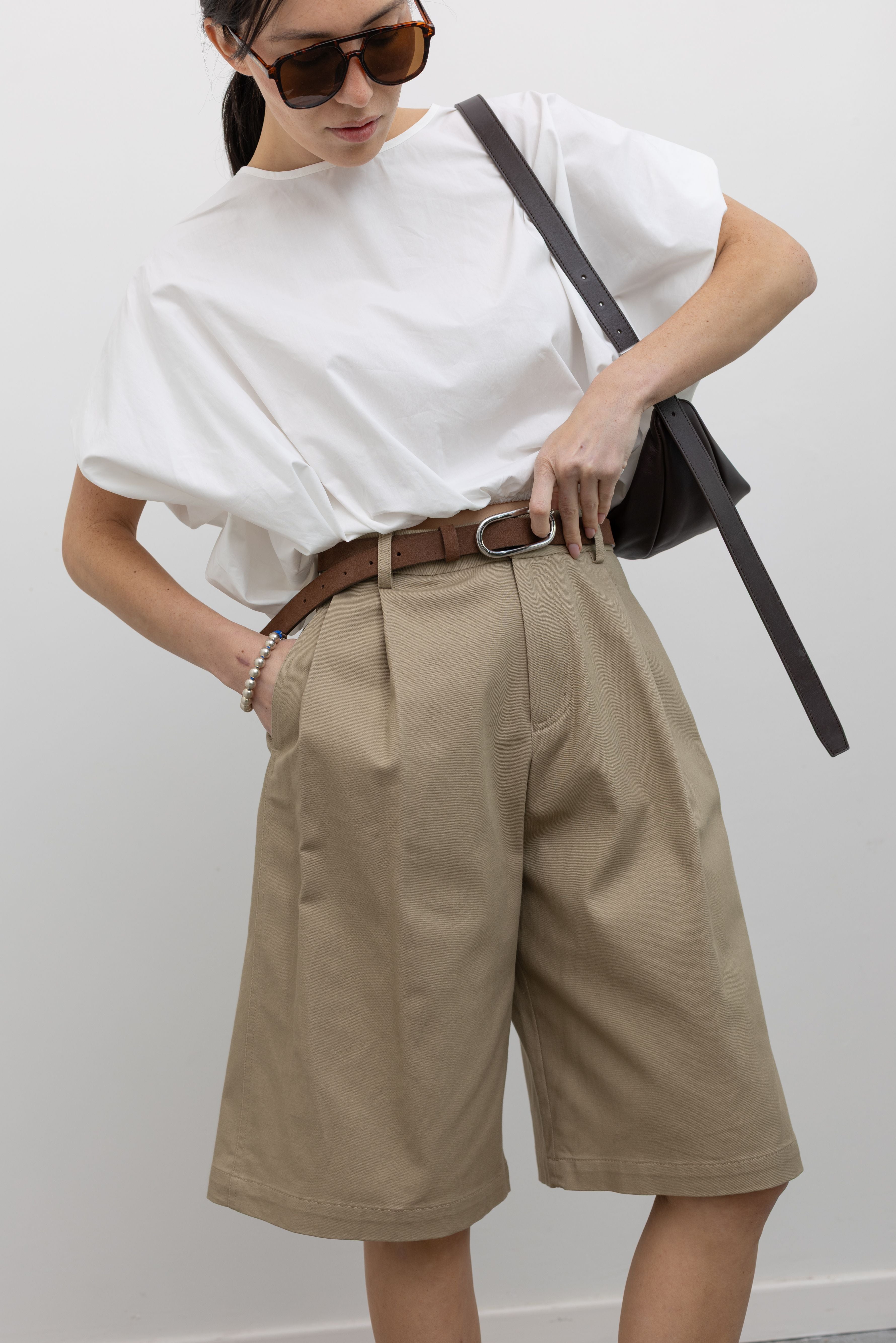Close-up front view of a woman posing wearing a 90's inspired wardrobe staple in the Tela sand cotton gabardine Bermuda long short featuring a front pleat and side seam pockets paired with a classic brown leather belt and a crisp white top at an Alas Eius store.