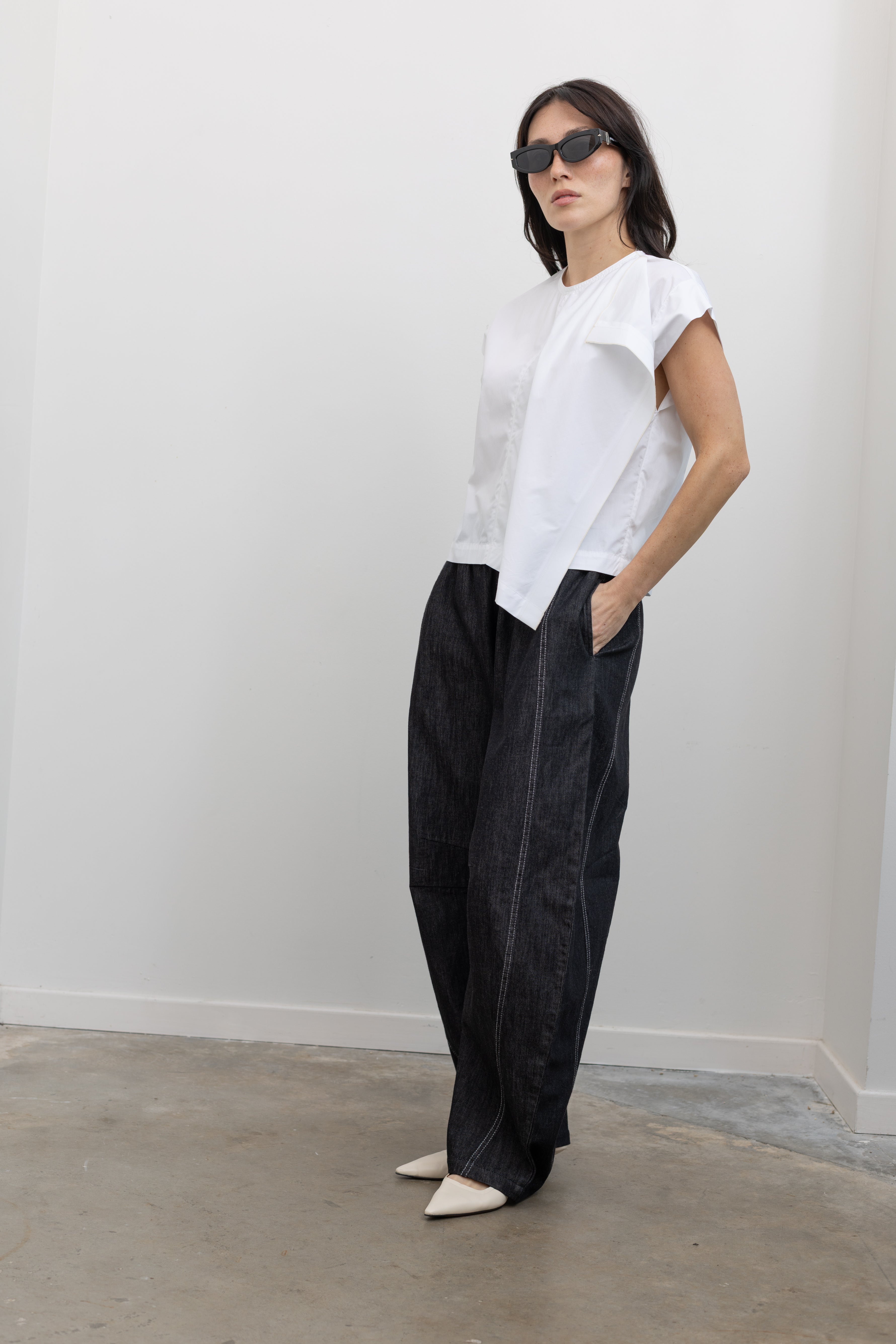 Full length 3/4 view of a woman wearing chic and modern spring fashion in a Tela brand white Livia shirt featuring a front pleat that creates an intentional asymmetrical drape, crew neckline and relaxed cap sleeves paired with oversized baggy barrel leg pants at at an Alas Eius store. 