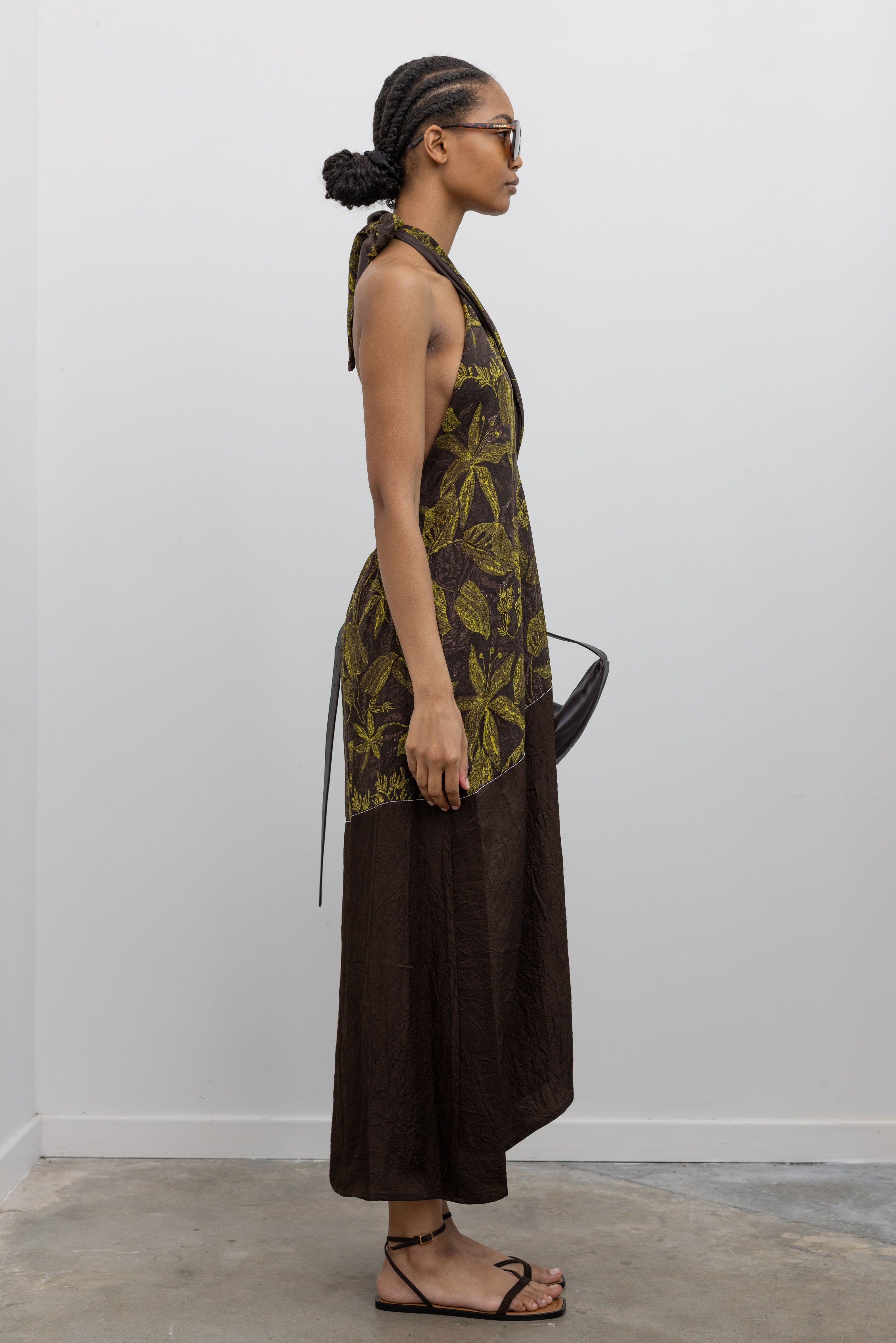 Full length side view of a woman standing wearing summer fashion in a Tela label olive green floral print halter neck midi dress with a contrasting chocolate brown hem paired with a St. Agni leather bag and strappy sandals at an Alas Eius Store.
