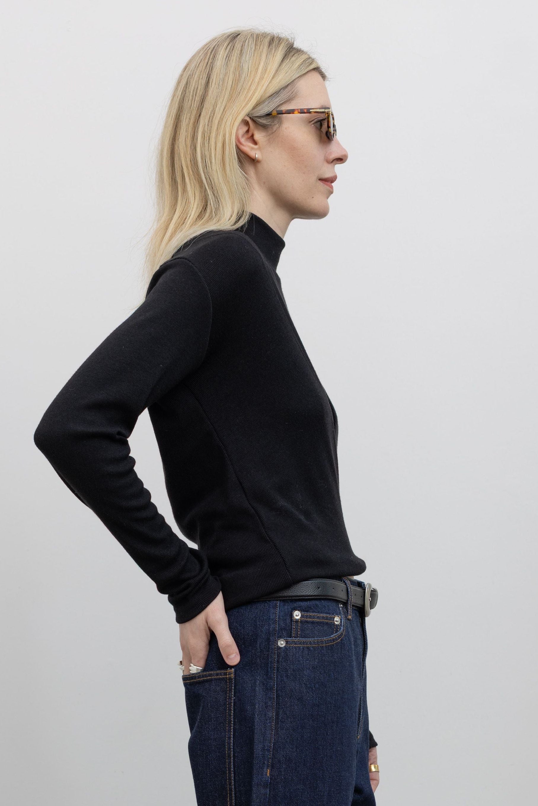 Side detail view of woman standing wearing Tibi Dark Wash Denim 5 Pocket Jeans with Alas Eius Label black mock neck sweater.