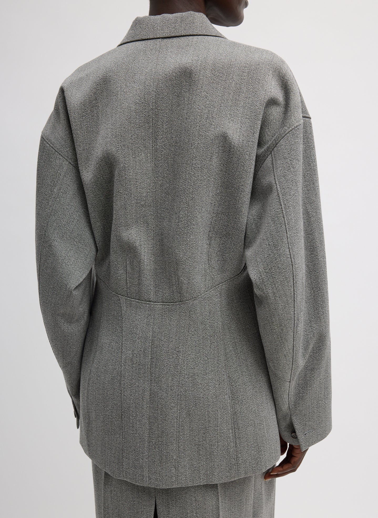 Close-up back view of a woman standing wearing sophisticated and modern spring fashion in the Tibi Light Gray Structured Wool Sculpted Blazer crafted from firm, refined virgin wool and features a blousant back with sculpted seaming and a button-front closure at an Alas Eius store. 