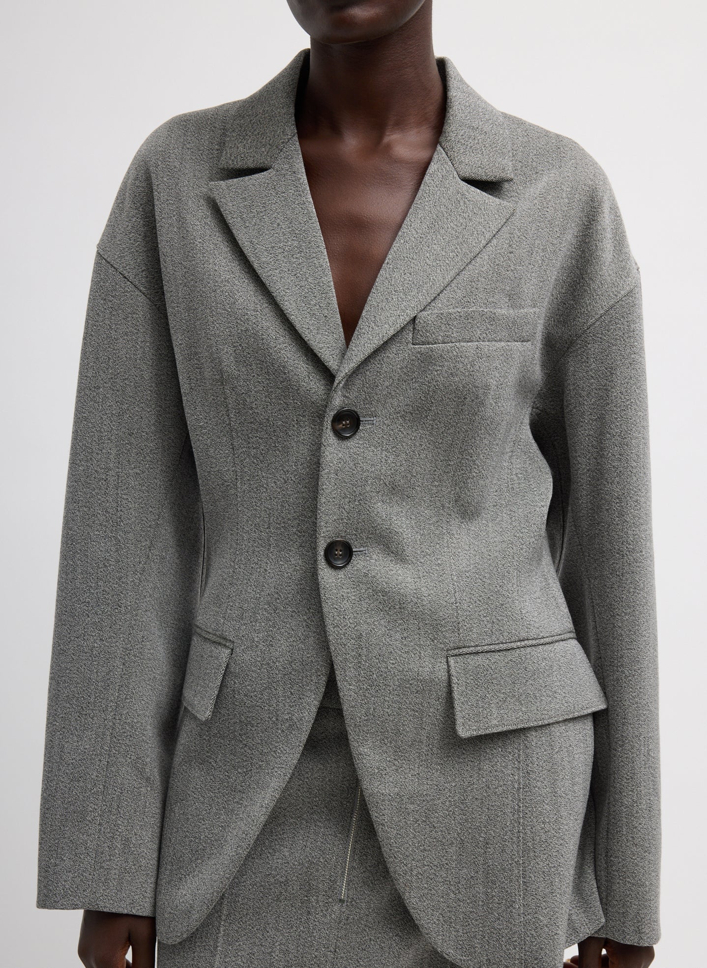 Close-up view of a woman standing wearing sophisticated and modern spring fashion in the Tibi Light Gray Structured Wool Sculpted Blazer crafted from firm, refined virgin wool and features a blousant back with sculpted seaming and a button-front closure at an Alas Eius store. 