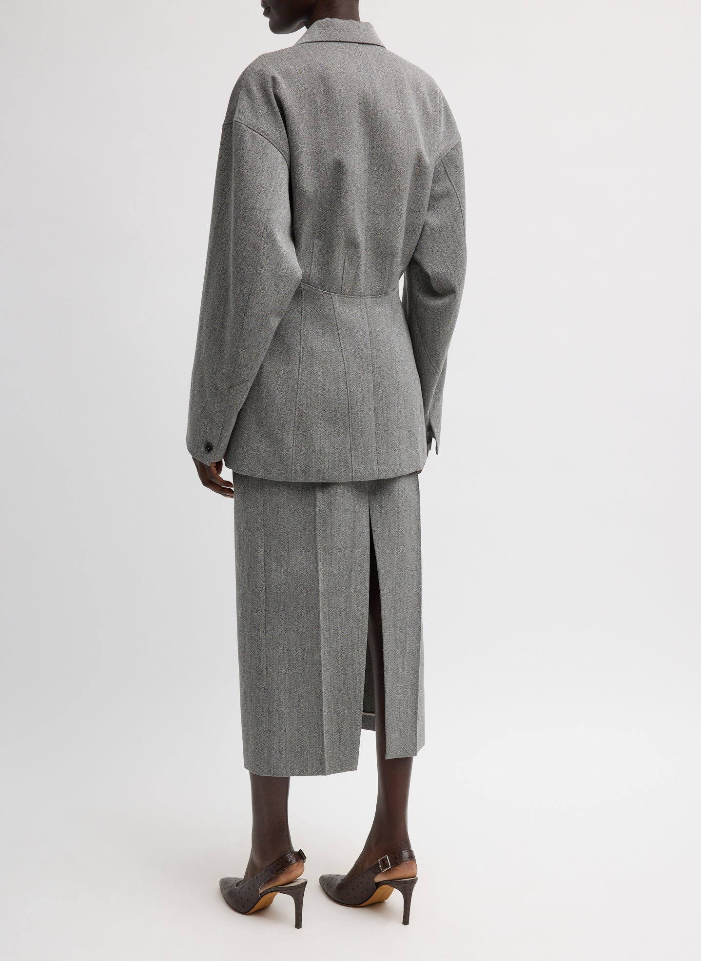 Back view of a woman standing wearing sophisticated and modern spring fashion in the Tibi Structured Wool Sculpted Blazer crafted from firm, refined virgin wool and features a blousant back with sculpted seaming and a button-front closure at an Alas Eius store. 