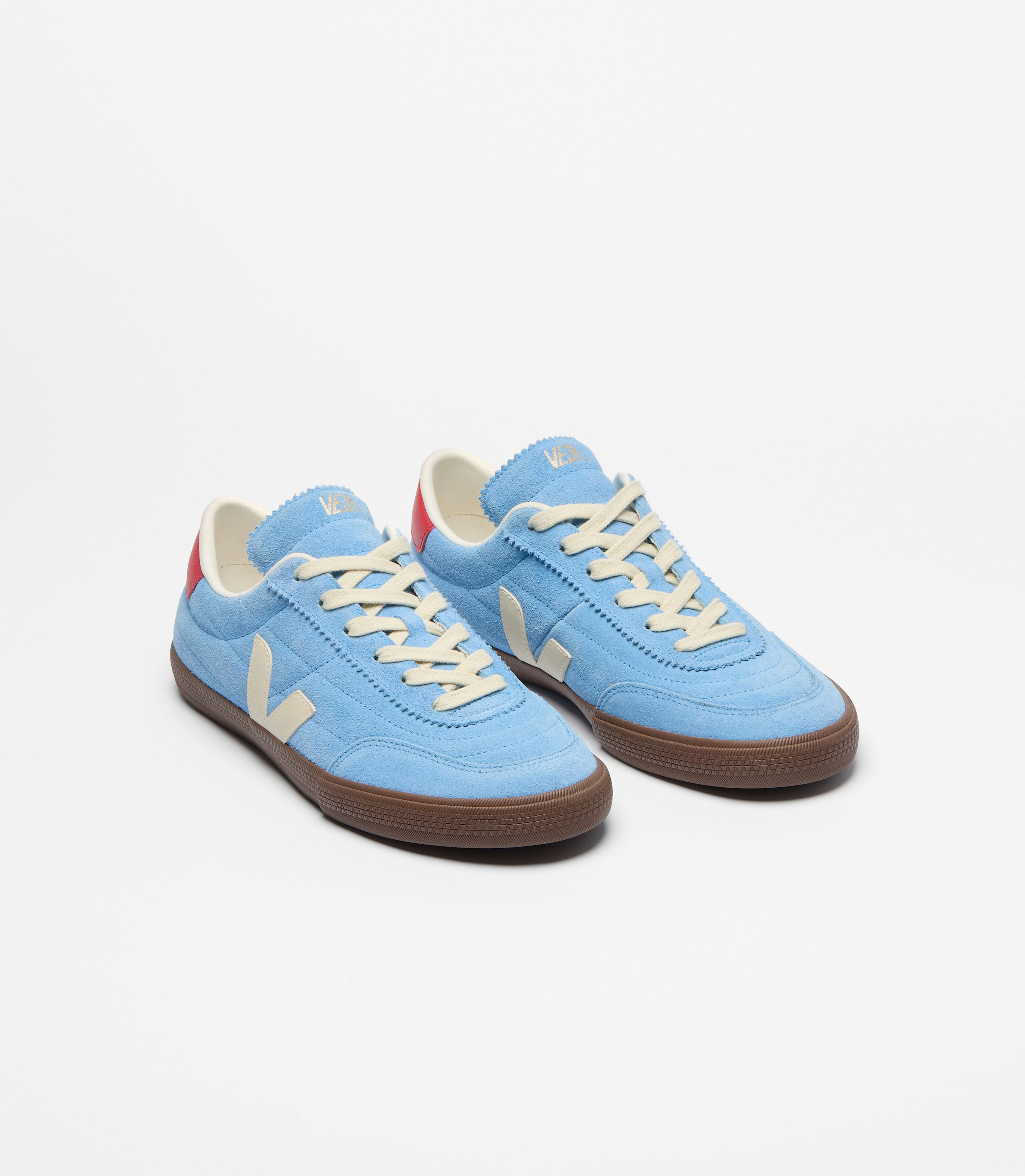 A pair of Veja sneakers in a sky blue suede upper paired with contrasting cream details and a rich brown sole. Finished with a bold red heel tab and signature side logo, the design blends sport and urban culture with modern everyday style at an Alas Eius store. 
