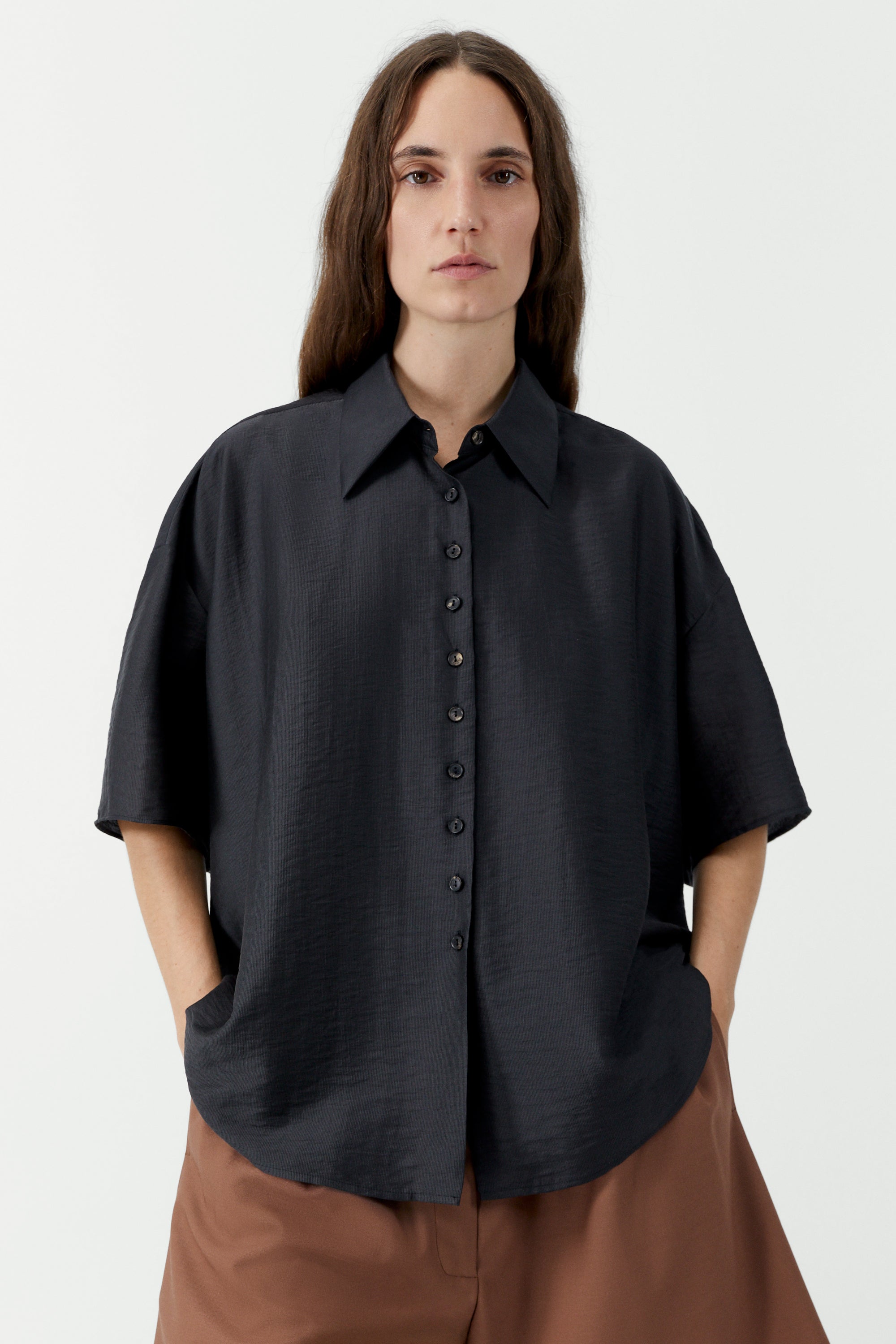 Woman standing wearing SS26 contemporary womenswear in the Cordera Espresso Brown Short Sleeve Button Front Shirt featuring an oversized, cocoon-shape, roomy short sleeves, pointy collar, small contrasting buttons down the front at an Alas Eius store. Made in Spain. 