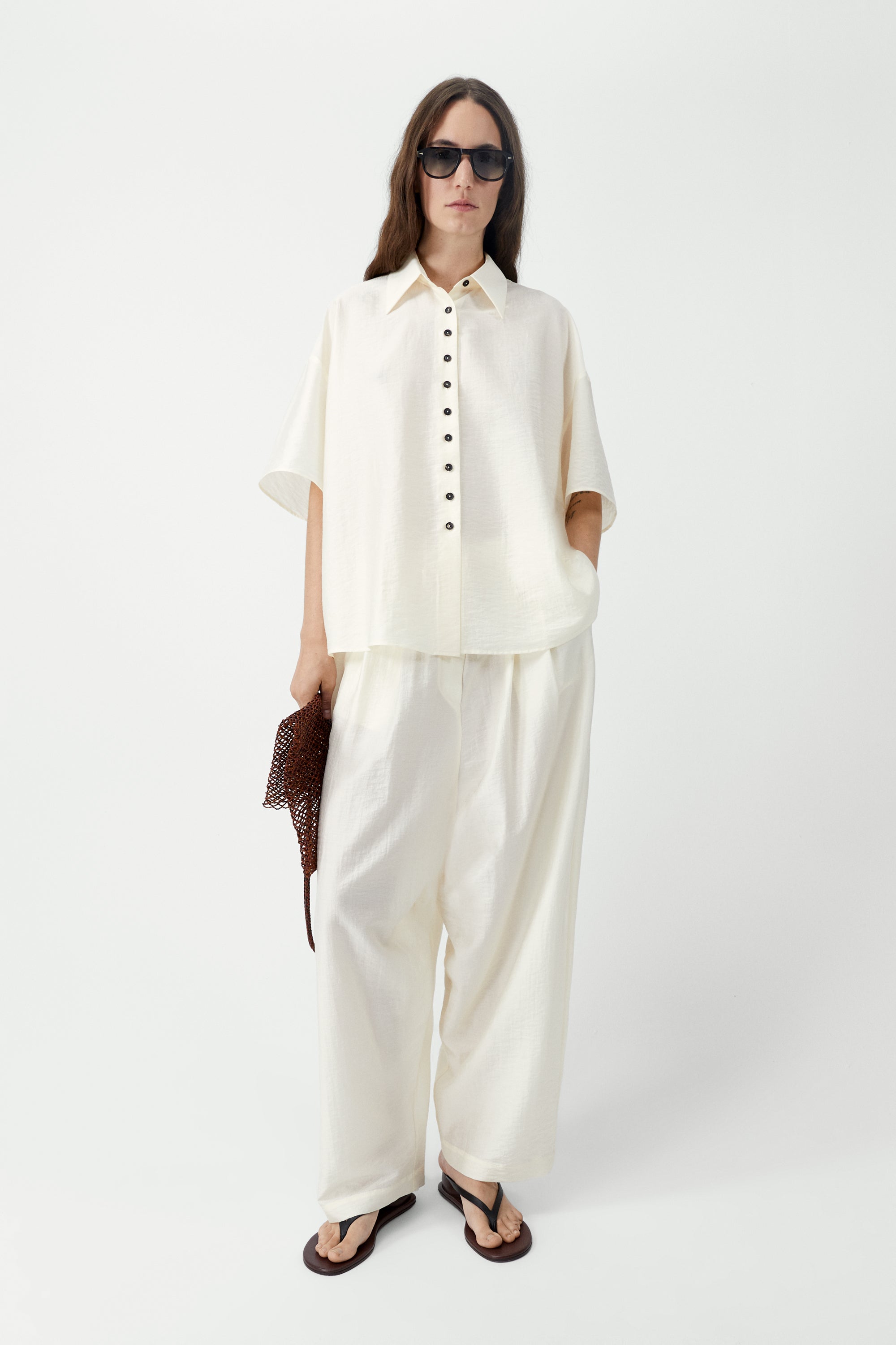 Full length front view of a woman standing wearing SS26 contemporary womenswear in the Cordera marfil Short Sleeve Button Front Shirt featuring an oversized, cocoon-shape, roomy short sleeves, pointy collar, small contrasting buttons down the front at an Alas Eius store. Made in Spain. 
