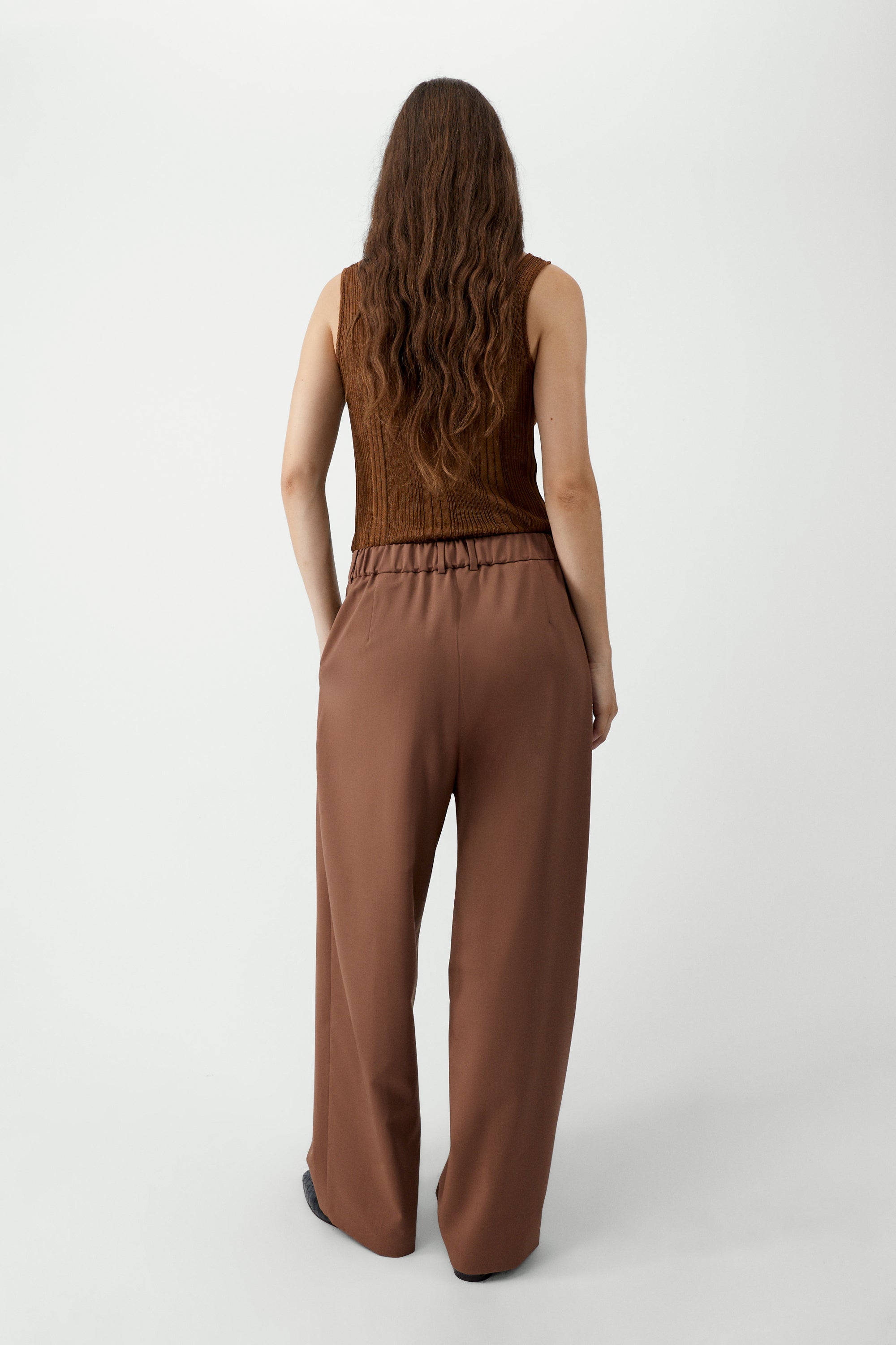 Back view of a woman wearing an elevated wardrobe staple in the Cordera Clove Tailored Masculine Pants featuring a loose, masculine fit with a mid-rise and double front pleats for added structure. Details include jetted side pockets, back welt pockets, an elasticated waistband at the back, zip and button closure, and belt loops at Alas Eius. 